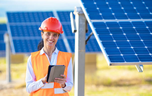 istock A female engineer or electrical technician wearing an orange vest and hard hat stands holding a notepad and smiling while working outdoors with the solar panel 1373564283