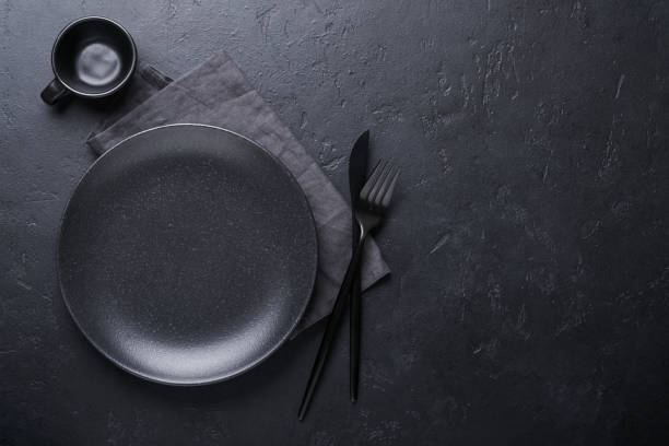 Black spoon, fork, knife and clean empty black plate. Set of stylish black and gold cutlery on black background. Fashionable and luxury eating. Top view. Copy space for your text. stock photo