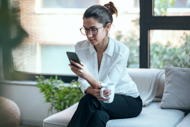 istock Pretty young woman using mobile phone while drinking cup of coffee on sofa at home. 1373614869
