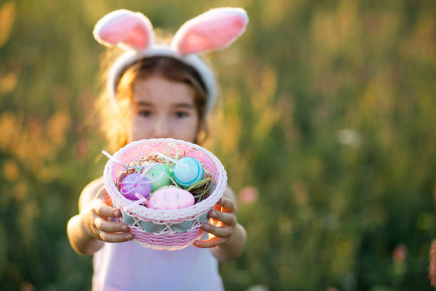 jolie fille drôle avec des œufs de pâques peints au printemps dans la nature dans un champ avec la lumière dorée du soleil et des fleurs. vacances de pâques, lapin de pâques avec oreilles, œufs colorés dans un panier. mode de vie - easter or easter bunny or easter egg or easter basket not business not silhouette photos et images de collection