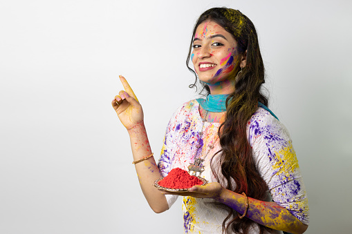 Young smart indian girl face coloured with gulal with plate full of color powder for festival of colours Holi, a popular hindu festival celebrated across india advertisement isolated white background
