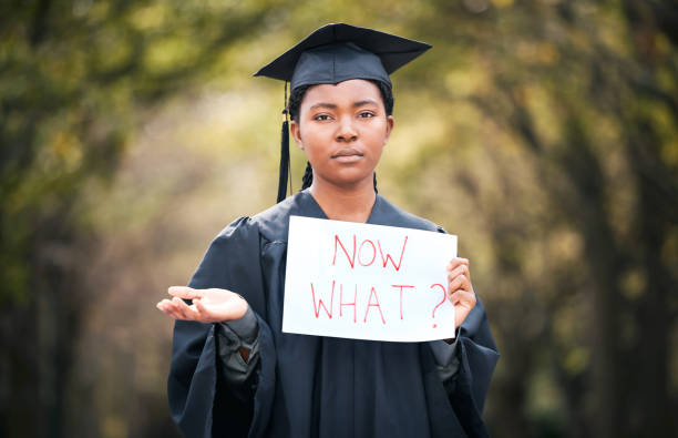 retrato de una mujer joven sosteniendo un letrero que dice "ahora qué" el día de la graduación - unemployment fotografías e imágenes de stock