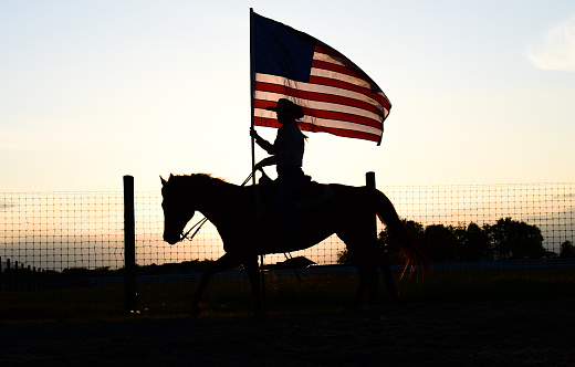 silhouette of a girl riding her horse in the sunset while flying the American flag