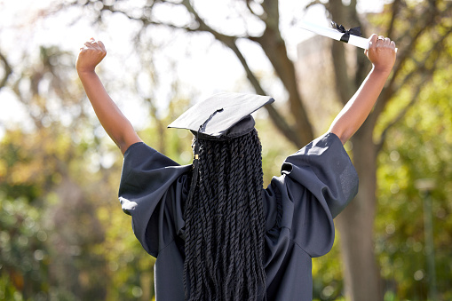 A female University graduate, of African decent, poses for a portrait outside on campus.  She is dressed formally in a gown and cap as she poses for a graduation photo.