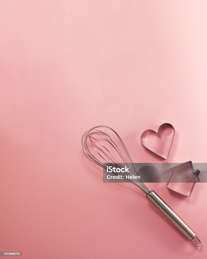 Steel whisk house and heart-shaped cutter flat lay top view. Confectionery cooking concept with copy space on bright pink paper background. National cooking day. Above Stock Photo