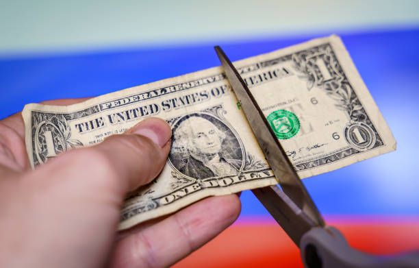 hand mit einer schere, die einen dollarschein mit der russischen flagge im hintergrund schneidet. - president of russia stock-fotos und bilder