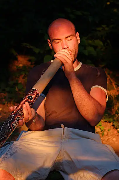 Young guy play on didgeridoo with the australian aboriginal musical instrument in lights of sunset