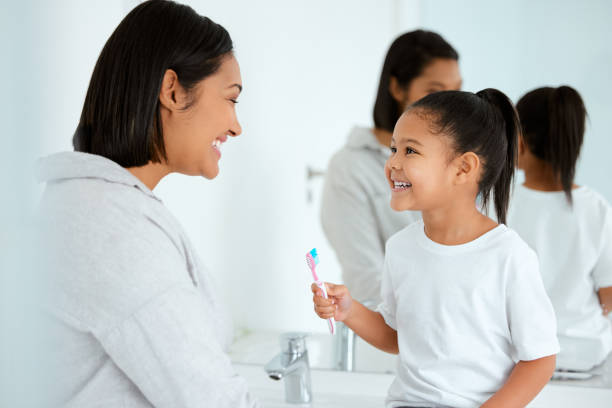 aufnahme eines entzückenden kleinen mädchens, das sich die zähne putzt, während ihre mutter ihr zu hause hilft - mirror mother bathroom daughter stock-fotos und bilder