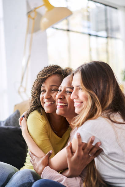 vertikales foto von drei glücklichen freundinnen, die sich lächelnd umarmen. lustige frauen feiern zusammen auf dem wohnzimmersofa - freundin stock-fotos und bilder