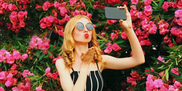 schöne frau, die selfie-foto per telefon macht, die ihre roten lippen bläst und süßen luftkuss auf rosa blumen sendet rosenhintergrund - fotografieren grafiken stock-fotos und bilder