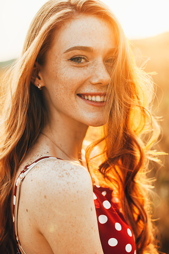 Close-up portrait of a red-haired romantic woman with freckled face enjoying nature, smiling and looking at camera. Sun light on sunset.