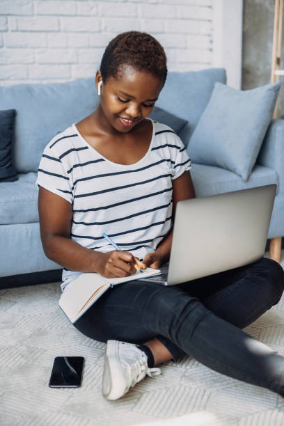 istock Afro woman studying remotely attending an online conference and taking notes in a notebook. Sitting on floor using her laptop. Internet technology. Online connection. Online education. 1374046917
