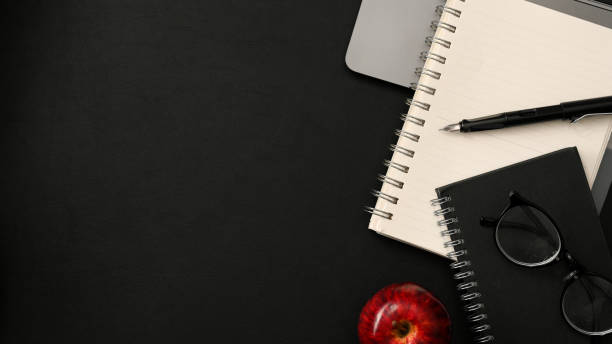 Overhead shot, Dark modern workspace with spiral notebooks, pen, eyeglasses, apple and copy space on black background.