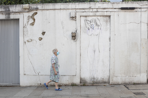 Older woman wearing a mask walks past  street art of a woman weeping in Santiago de Compostela.