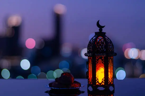 Selective focus of lantern that have moon symbol on top and small plate of dates fruit with city bokeh light background for the Muslim feast of the holy month of Ramadan Kareem.