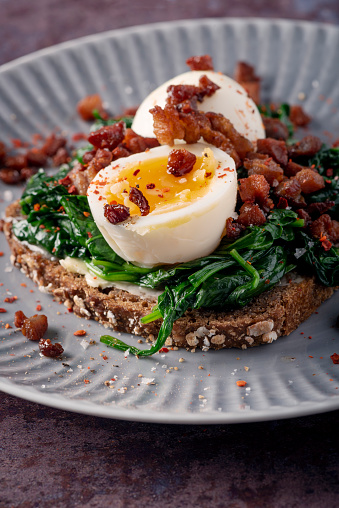 An open-faced sandwich  or in danish “smørrebrød”. Made with buttered rye bread with wilted spinach, pancetta and topped off with a soft boiled egg and fresh garden cress. Colour, vertical format with some copy space. Smorrebrod are a staple of the danish food culture, a very popular choice for lunch or a light dinner. Toppings can be almost anything from roast beef to prawns but the rye bread at the bottom of the sandwich has to be good!