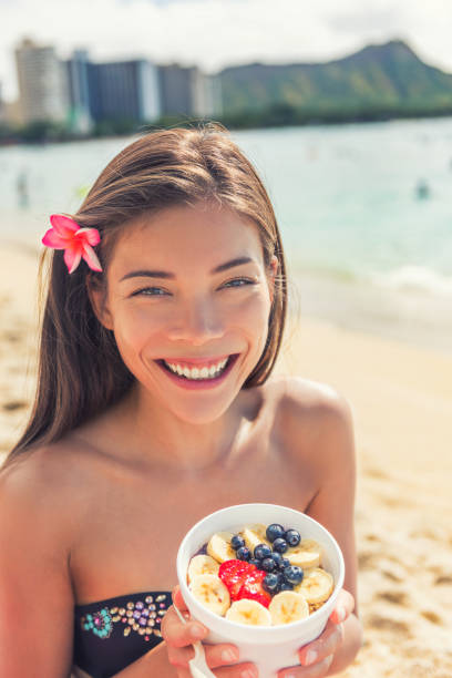 açaí tigela de café da manhã saudável mulher asiática comendo lanche no fundo do oceano na praia do havaí. frutas e frutas frescas ao ar livre para uma dieta de perda de peso. comendo prato havaiano local - outdoors waikiki waikiki beach honolulu - fotografias e filmes do acervo