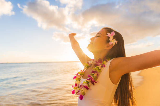 ハワイの休暇で自由に両手を広げて踊るワイキキビーチでハワイアンレイフラワーネックレスを身に着けているハワイフラルアウの女性.新鮮な花の髪を持つアジアの女の子、伝統的なポリネ - hula dancing ストックフォトと画像