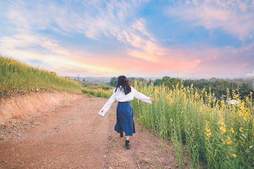 Asian woman in a white shirt walks on a dirt road in the middle of a field of bright yellow flowers.