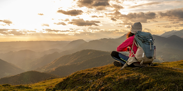 Rear view of woman sitting on top of mountain against cloudy sky during sunrise.