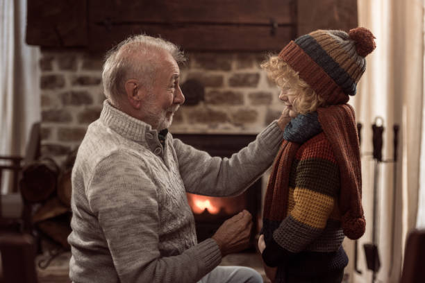 Grandfather putting winter cap on grandson's head in front of the fireplace because it's cold outside