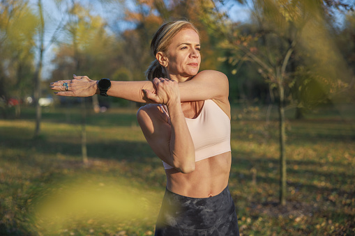 Mid adult woman wearing sports bra and leggings, stretching arm muscles and looking at distance, relaxing after workout in nature