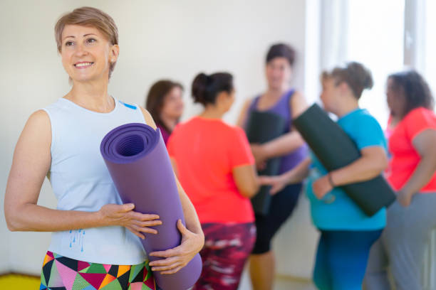 ladies holding yoga mats chatting before a class, one lady in the foreground is looking into the distance and smiling - teen obesity imagens e fotografias de stock