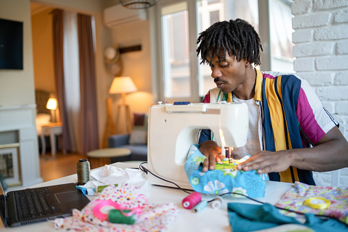 Using some brightly colored fabric, an Afro-Caribbean man is guiding the material while he stitches it using a sewing machine, more fabric and sewing implements covering the table he is working at at home on his dining table