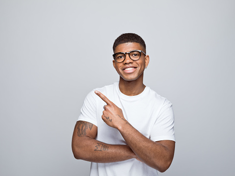 Happy african young man wearing white t-shirt and eyeglasses, pointing with index finger at copy space and smiling at camera. Studio portrait on grey background.