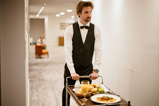 Waiter in uniform delivering tray with food in a room of hotel. Room service.