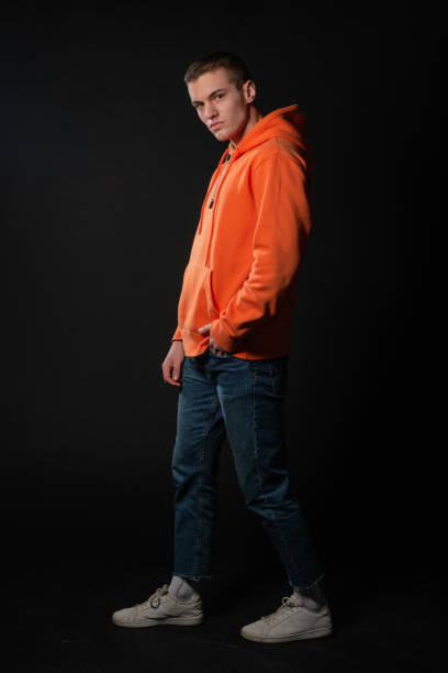 istock Young handsome man in jeans and orange hoodie posing while standing on a black background. 1374524675