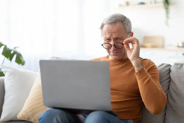 Poor Eyesight. Senior Man Squinting Eyes Using Laptop Wearing Eyeglasses Having Problems With Vision Sitting On Couch. Ophtalmic Issue, Bad Sight In Older Age, Macular Degeneration Concept