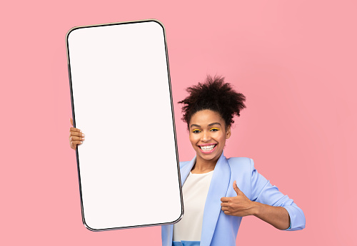 Happy glad excited young african american female blogger in suit show thumb up gesture and huge phone with empty screen, on pink background, cut out. Great news, app, ad, device and recommendation