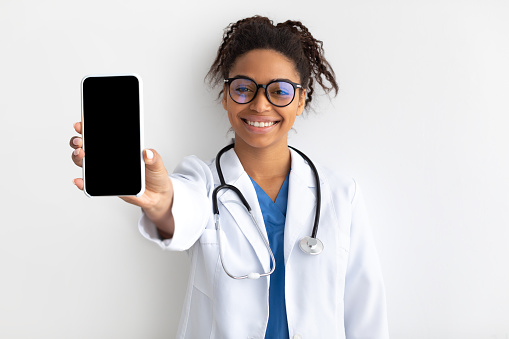 Smiling Black Female Physician In Eyeglasses And Uniform Showing Blank Cell Phone Screen Close To Camera Holding Gadget In Hand Isolated On White Studio Background, Mock Up. Telemedicine Concept