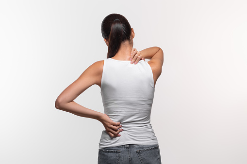 Unrecognizable long-haired brunette woman with neck and back pain rubbing her painful body, lady suffering from office syndrome, back view, white studio background, panorama with copy space