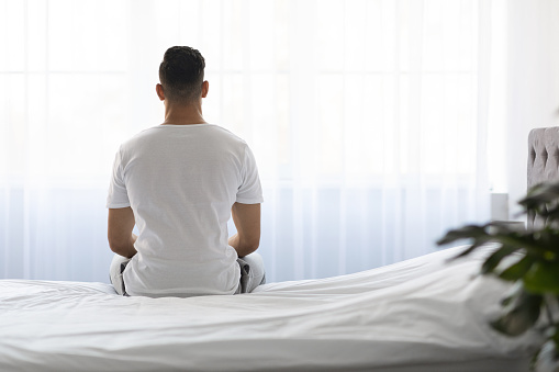 Loneliness Concept. Rear View Of Young Man Sitting On Bed And Looking At Window In The Morning, Unrecognizable Male In White T-Shirt Spending Time In Light Bedroom After Waking Up, Copy Space