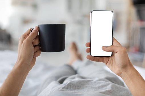 Male Hands Holding Smartphone With Blank Screen And Cup Of Coffee While Lying In Bed At Home, Unrecognizable Man Using Modern Gadget With Copy Space For Website Or Mobile App Design, Mockup