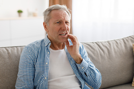 Dental Care, Tooth Sensitivity Concept. Portrait of unhappy mature man suffering from terrible toothache, touching cheek feeling acute pain, aged male sitting on the couch at home, free copy space
