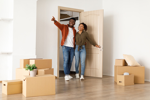 Real Estate Ownership. Excited Black Spouses Entering Their New Home, Husband Pointing Aside Showing Empty Living Room With Moving Boxes After Relocation. Family Housing Concept. Full Length