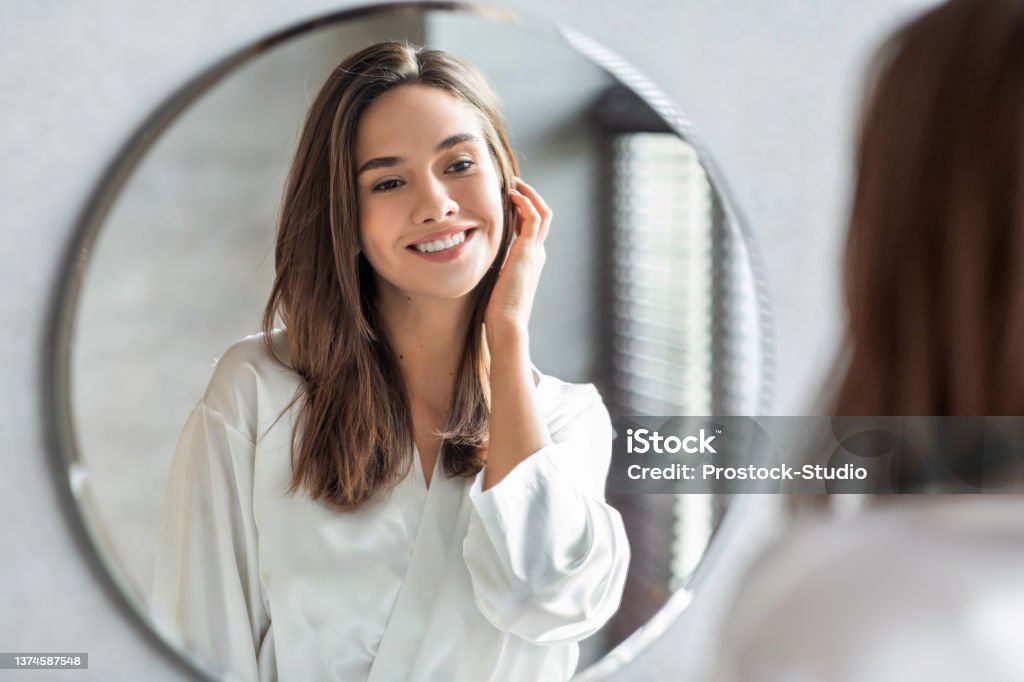 Beauty Concept. Portrait Of Attractive Happy Woman Looking At Mirror In Bathroom Beauty Concept. Portrait Of Attractive Happy Woman Looking At Mirror In Bathroom, Beautiful Millennial Lady Wearing White Silk Robe Smiling To Reflection, Enjoying Her Appearance, Selective Focus Women Stock Photo