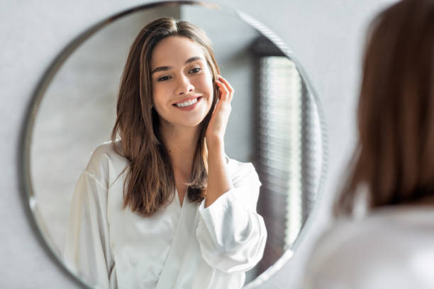 Beauty Concept. Portrait Of Attractive Happy Woman Looking At Mirror In Bathroom Beauty Concept. Portrait Of Attractive Happy Woman Looking At Mirror In Bathroom, Beautiful Millennial Lady Wearing White Silk Robe Smiling To Reflection, Enjoying Her Appearance, Selective Focus human face stock pictures, royalty-free photos & images