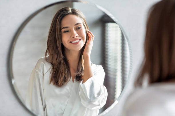 Beauty Concept. Portrait Of Attractive Happy Woman Looking At Mirror In Bathroom Beauty Concept. Portrait Of Attractive Happy Woman Looking At Mirror In Bathroom, Beautiful Millennial Lady Wearing White Silk Robe Smiling To Reflection, Enjoying Her Appearance, Selective Focus fresh faced stock pictures, royalty-free photos & images