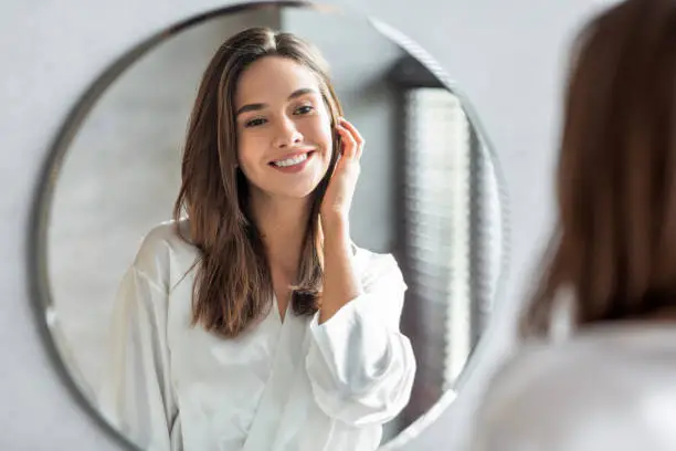 Beauty Concept. Portrait Of Attractive Happy Woman Looking At Mirror In Bathroom, Beautiful Millennial Lady Wearing White Silk Robe Smiling To Reflection, Enjoying Her Appearance, Selective Focus