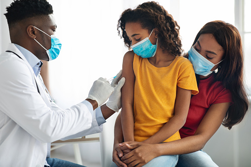 African american doctor making vaccination against coronavirus in shoulder for child, african american family in protective face masks visiting pediatrician at clinic. Kids immunization concept