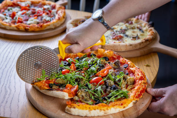 istock Cutting pizza with vegetables and arugula served on round wooden board, using cutter 1374648005