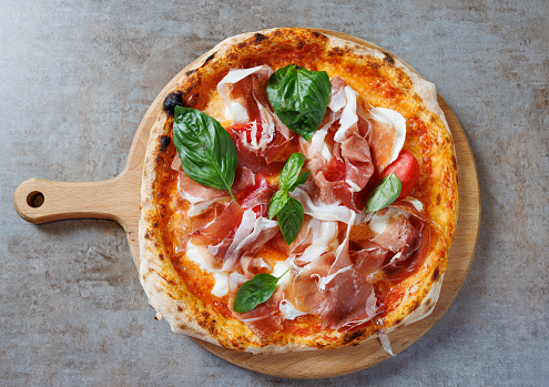 Pizza garnished with prosciutto, basil and cherry tomato, cheese and tomato sauce base, served on round wooden board