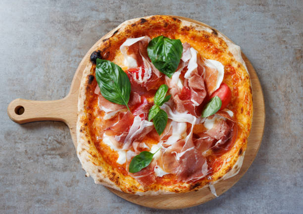 Pizza with prosciutto and basil served on round wooden board Pizza garnished with prosciutto, basil and cherry tomato, cheese and tomato sauce base, served on round wooden board flatbread photos stock pictures, royalty-free photos & images