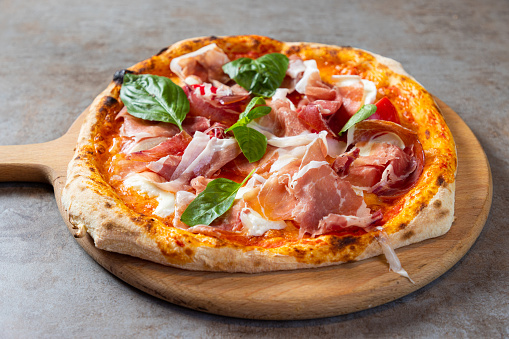 Close-up on pizza with tomato sauce and cheese base, garnished with prosciutto, basil and cherry tomato served on round wooden board