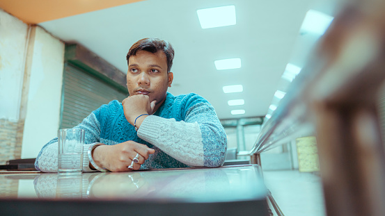 In this indoor image with copy space, an Asian/Indian young man contemplates sitting in a cafeteria.