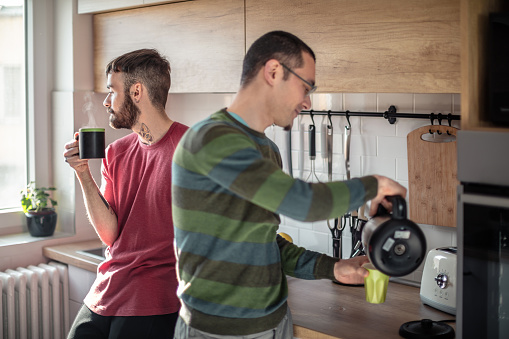 Gay couple together in the kitchen, bearded young man is holding a cup of coffee and looking out the window, his partner holding kettle and pouring fresh coffee for himself
